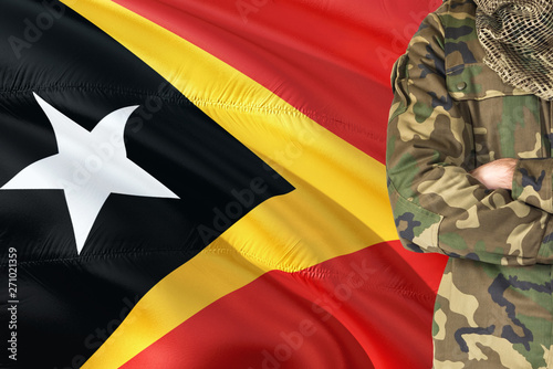 Crossed arms Timorese soldier with national waving flag on background - East Timor Military theme.