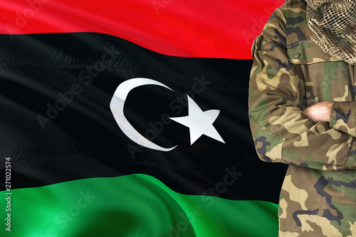 Crossed arms Libyan soldier with national waving flag on background - Libya Military theme.