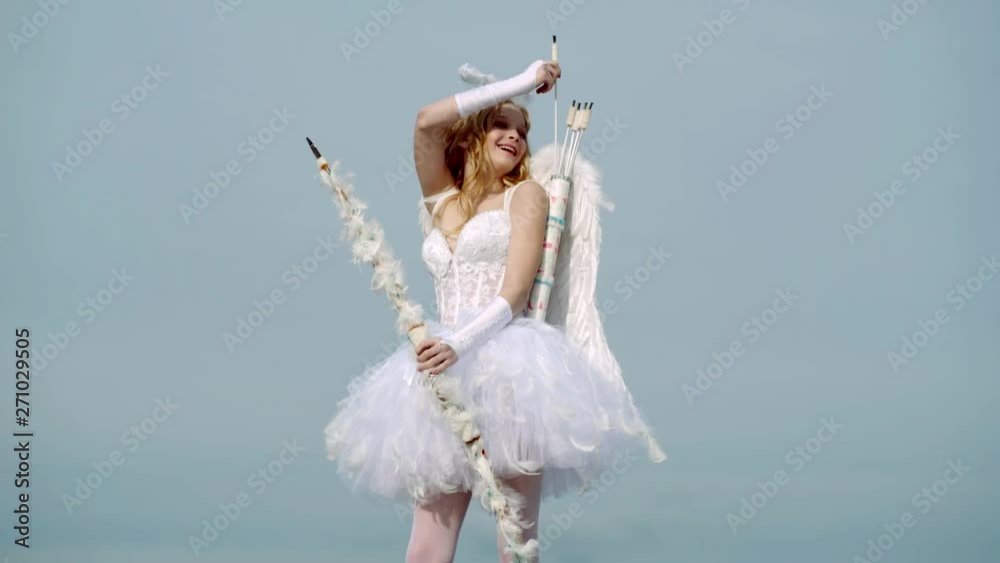 Concept of Valentines Daygirl dressed as an angel on a light background. arrow and wings. Innocent Girl with angel wings standing with bow and arrow against blue sky and white clouds
