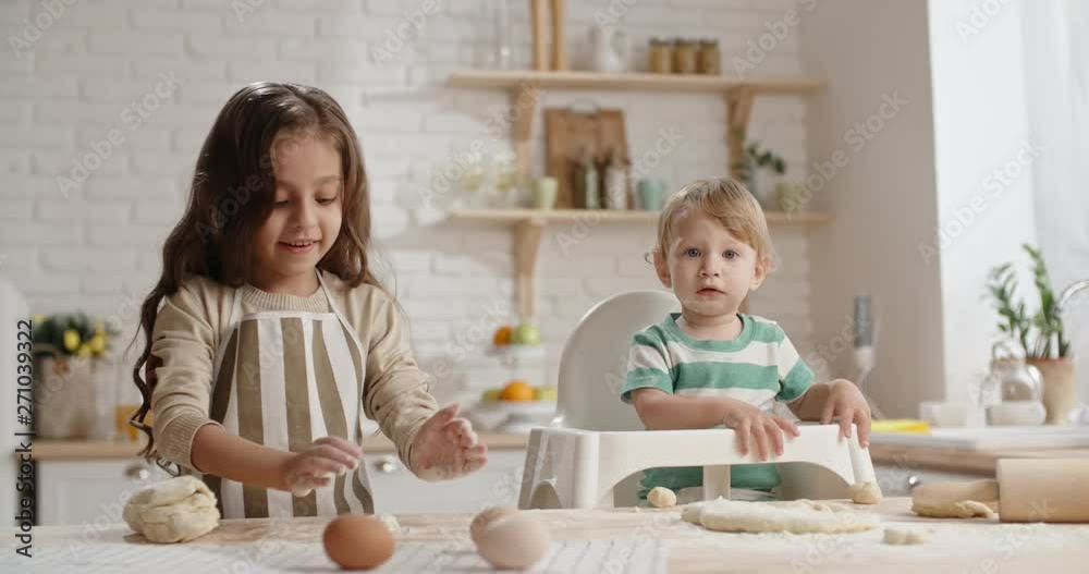 Little girl is learning to cook, preparing cake dough while her little brother in baby chair with floured face is playing with piece of pastry - togetherness, childhood dream 4k Little girl is learning to cook, preparing cake dough while her little brother in baby chair with floured face is playing with piece of pastry - togetherness, childhood dream 4k