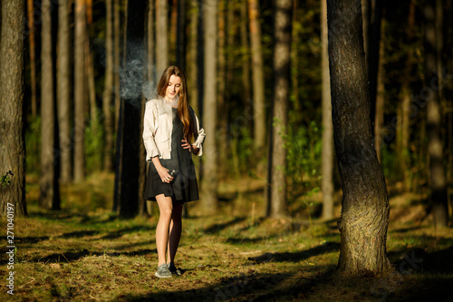 Vape teenager. Young cute girl in casual clothes smokes an electronic cigarette outdoors in the forest at sunset in summer. Bad habit that is harmful to health. Vaping activity.