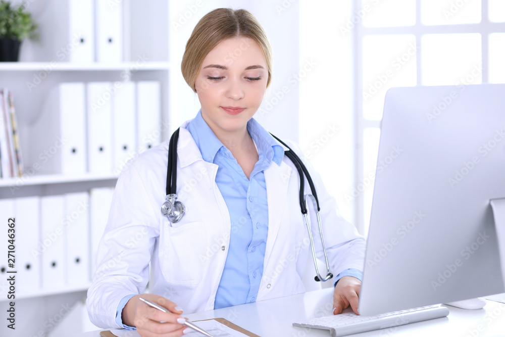 Young woman doctor at work in hospital looking at desktop pc monitor. Physician controls medication history records and exam results. Medicine and healthcare concept Young woman doctor at work in hospital looking at desktop pc monitor. Physician controls medication history records and exam results. Medicine and healthcare concept