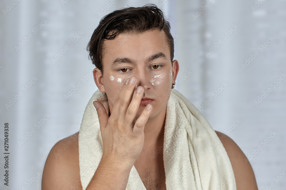 Young handsome caucasian man applying cream under the eyes, towel on shoulders. Caring face, metrosexual daily routine in the bathroom. Indoors, copy space. Young handsome caucasian man applying cream under the eyes, towel on shoulders. Caring face, metrosexual daily routine in the bathroom. Indoors, copy space.