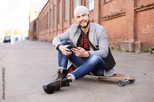 Handsome young man in grey coat and hat sitting on the longboard on the street in the city. Urban skateboarding concept. © ty