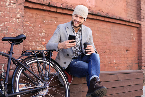 Handsome young man in grey coat and hat talking on the mobile phone and smiling while sitting near his bicycle outdoors.