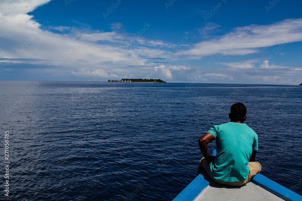 Man sitting on a traditional wooden boat heading over the calm tropical ocean towards a luxury resort on the atoll of the Maldives