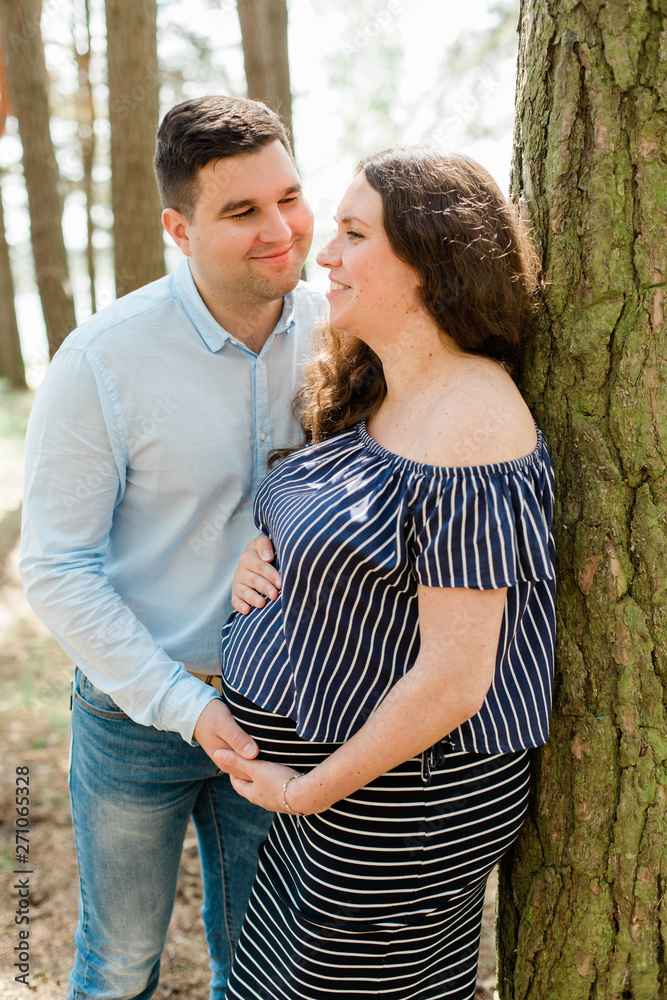Naklejka premium Cheerful pregnant woman with long hair standing on the beach with her husband behind. Young family expecting a child. Happy pregnancy concept.