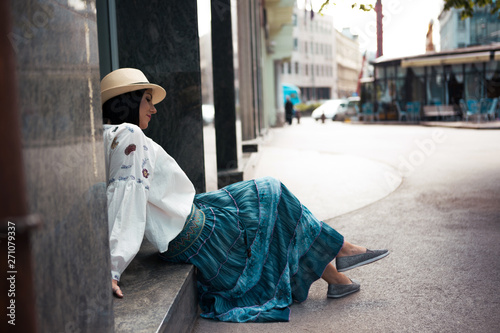 A young woman of black hair sits on the street, dressed in a gypsy style.