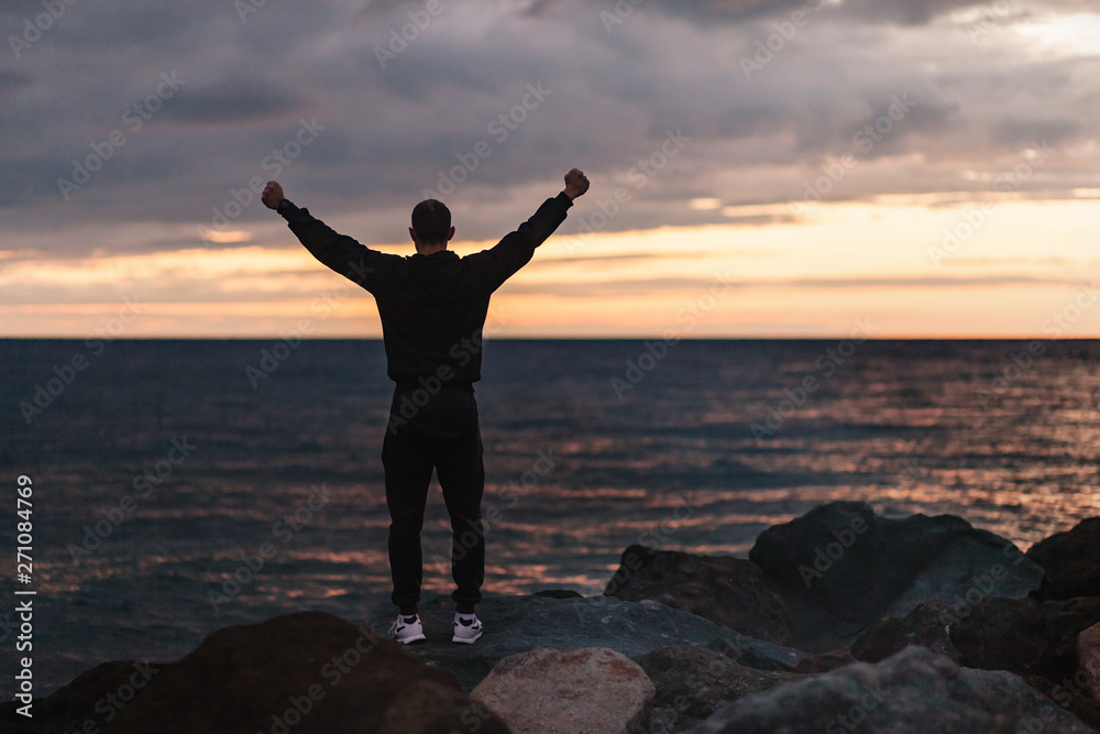 Rear view of a happy unidentified male athlete rejoices in a fulfilled dream by raising his hands up and looking at sunset by the sea on warm summer evening. Concept of achieving goals and results