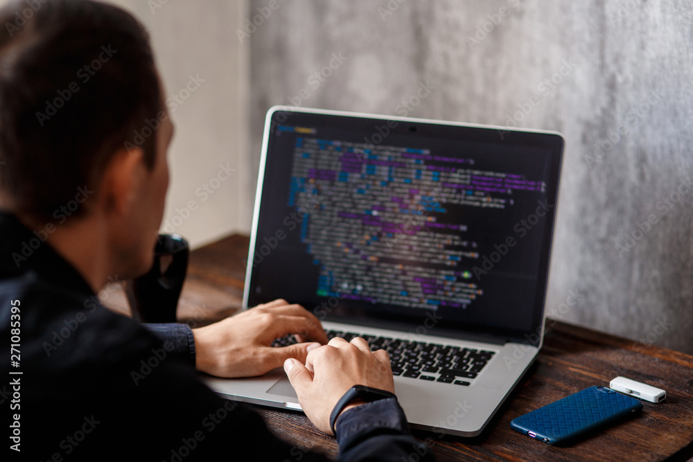 a male programmer in a black shirt is working at a laptop, close-up of the program code and developer's hands on a laptop. modern young programmer sitting at a wooden table at a computer a male programmer in a black shirt is working at a laptop, close-up of the program code and developer's hands on a laptop. modern young programmer sitting at a wooden table at a computer