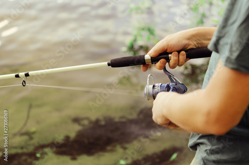 A man in a t-shirt and dark pants is fishing on the lake with a white fishing rod against the background of bushes and grass. Concept active leisure.