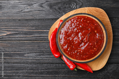 Bowl of hot chili sauce with red peppers on dark wooden background, top view. Space for text