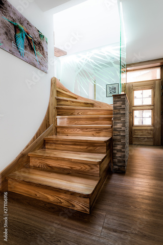 Wooden stairs with visible wooden texture in bright daylight, which is coming from the second floor. Stairs are lined with paneled stone wall and illuminated glass panel with a light blue touch.