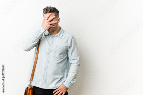 Young handsome business man blink at the camera through fingers, embarrassed covering face.