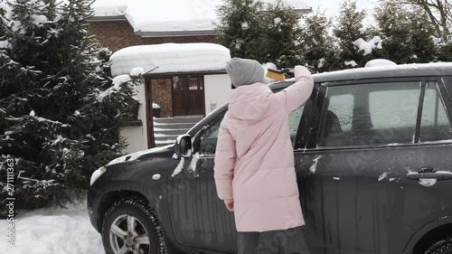 Young happy blonde woman in pink parka grey knit hat cleaning big black suv car from snow and ice, holding scraper brush tool in front of her house. Safe transportation winter driving concept.