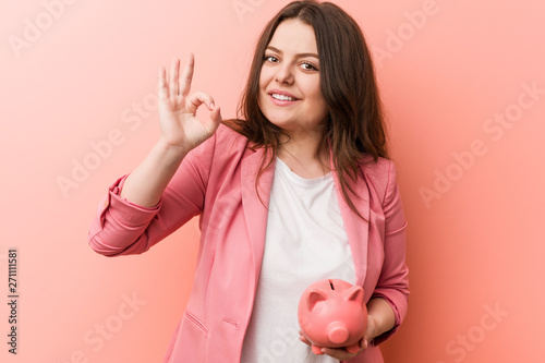 Young plus size curvy business woman holding a piggy bank cheerful and confident showing ok gesture.