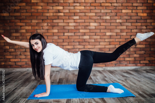 Attractive cheerful young woman working out indoors. Beautiful brunette model doing exercises on blue mat in room with walls.