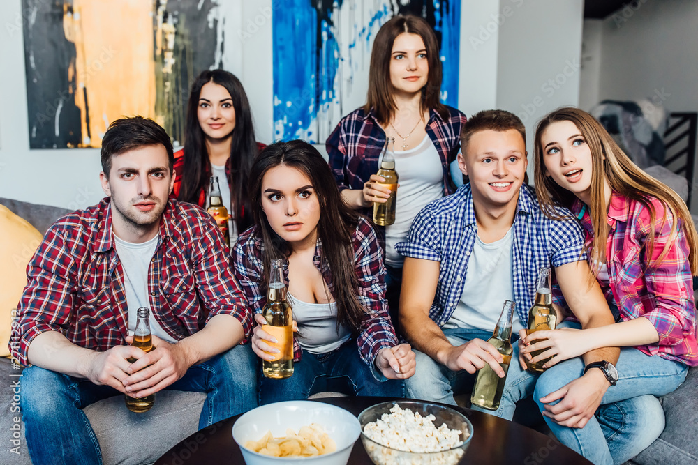 Young people having day-off relaxing in front of tv in cozy living room. Six friends spending their weekend together sitting on sofa, eating popcorn and laughing over extremely amusing tv-show.