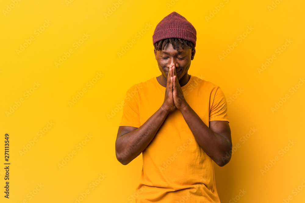 Young black man wearing rastas over yellow background holding hands in pray near mouth, feels confident. Young black man wearing rastas over yellow background holding hands in pray near mouth, feels confident.