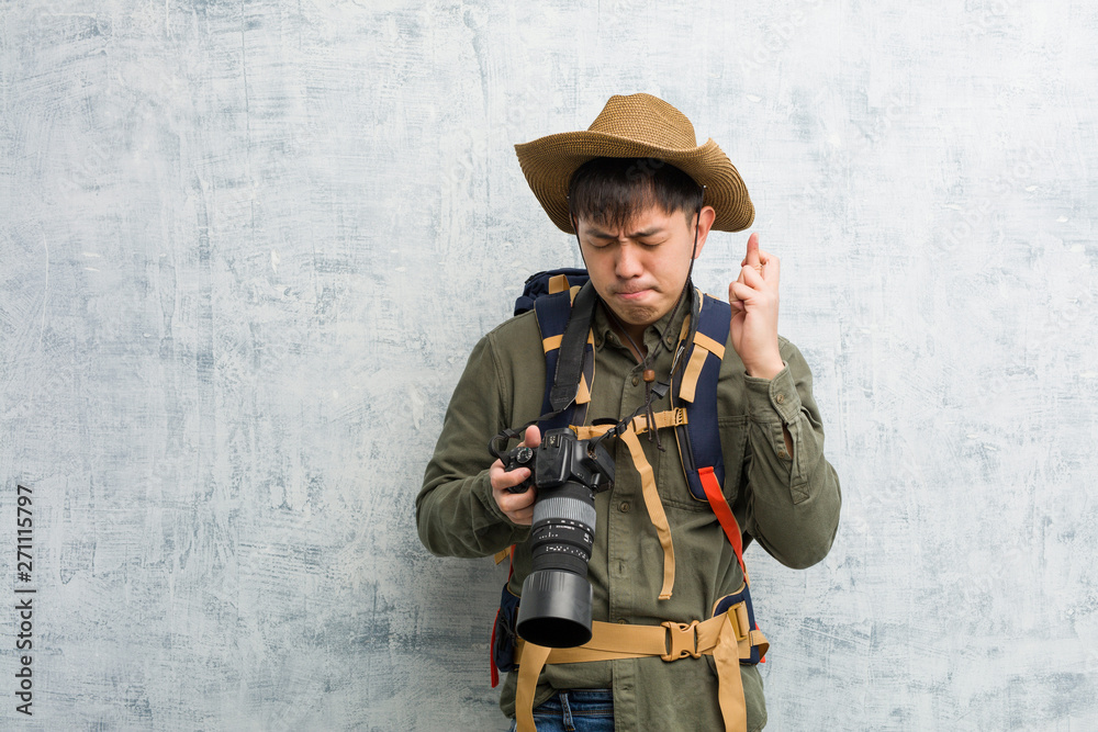Fototapeta premium Young chinese explorer man holding a camera crossing fingers for having luck