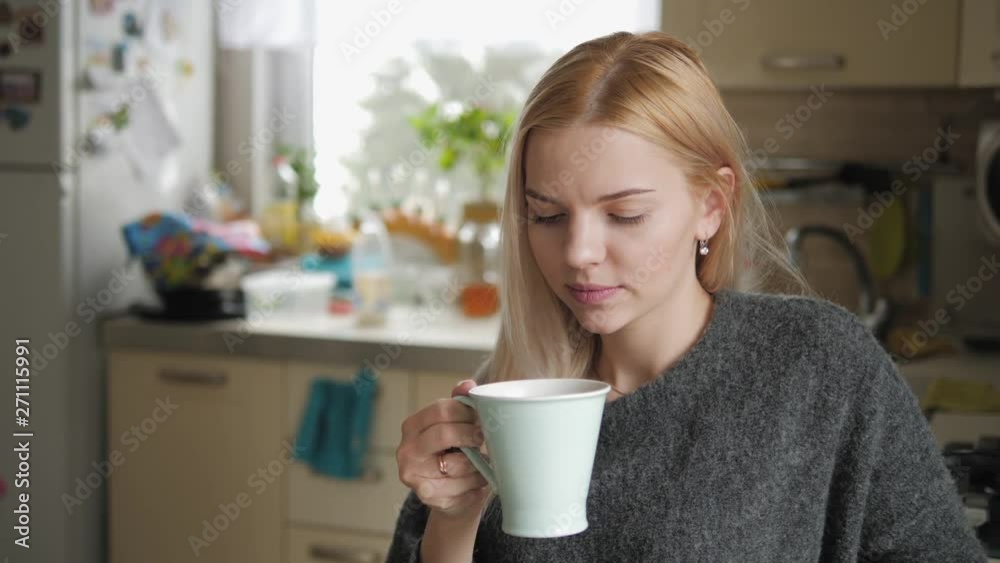 Medium closeup shot of young smiling adorable blonde european woman in grey sweater with mug posing against blurred cozy domestic kitchen interior background, looking out the window, smiling, laughing Medium closeup shot of young smiling adorable blonde european woman in grey sweater with mug posing against blurred cozy domestic kitchen interior background, looking out the window, smiling, laughing