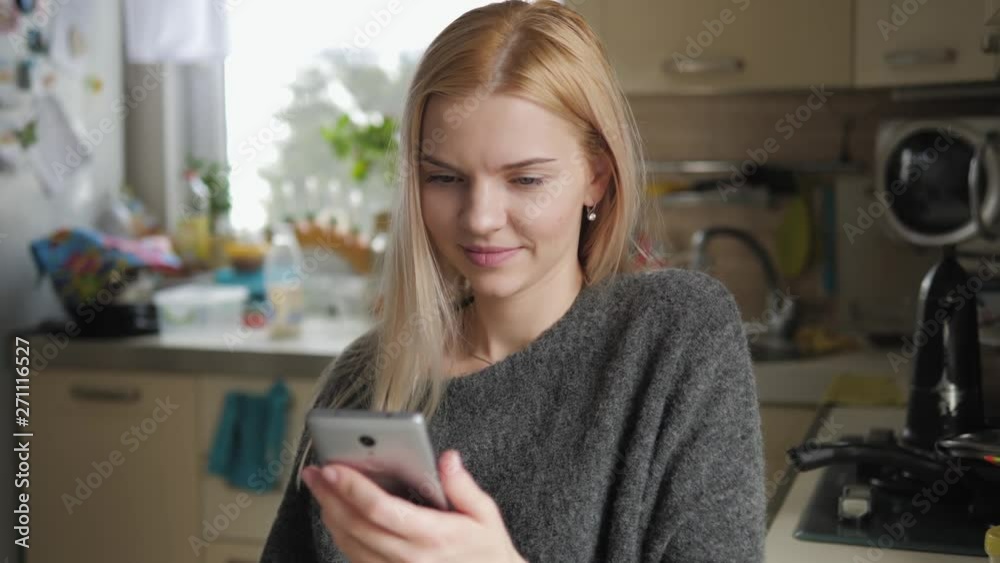 Medium closeup shot of happy fresh looking face young caucasian blonde woman using smartphone on domestic kitchen. Young attractive girl indoors using social networking app, scrolling, typing, swiping Medium closeup shot of happy fresh looking face young caucasian blonde woman using smartphone on domestic kitchen. Young attractive girl indoors using social networking app, scrolling, typing, swiping