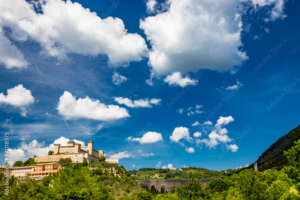View of Spoleto, green mountains, blue sky with white clouds. The Rocca Albornoziana fortress illuminated by the sun in summer. The bridge of the towers, Roman aqueduct. Trees in the foreground View of Spoleto, green mountains, blue sky with white clouds. The Rocca Albornoziana fortress illuminated by the sun in summer. The bridge of the towers, Roman aqueduct. Trees in the foreground