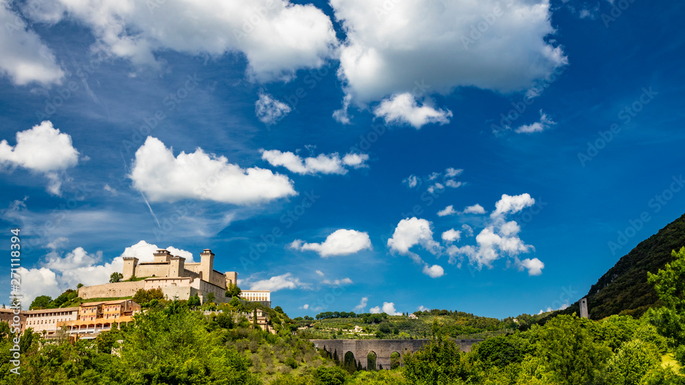 View of Spoleto, green mountains, blue sky with white clouds. The Rocca Albornoziana fortress illuminated by the sun in summer. The bridge of the towers, Roman aqueduct. Trees in the foreground