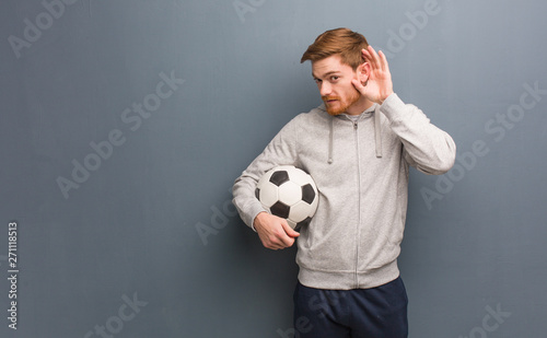 Young redhead fitness man try to listening a gossip. He is holding a soccer ball.