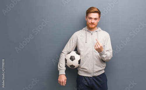 Young redhead fitness man inviting to come. He is holding a soccer ball.