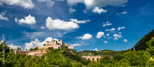 View of Spoleto, green mountains, blue sky with white clouds. The Rocca Albornoziana fortress illuminated by the sun in summer. The bridge of the towers, Roman aqueduct. Trees in the foreground