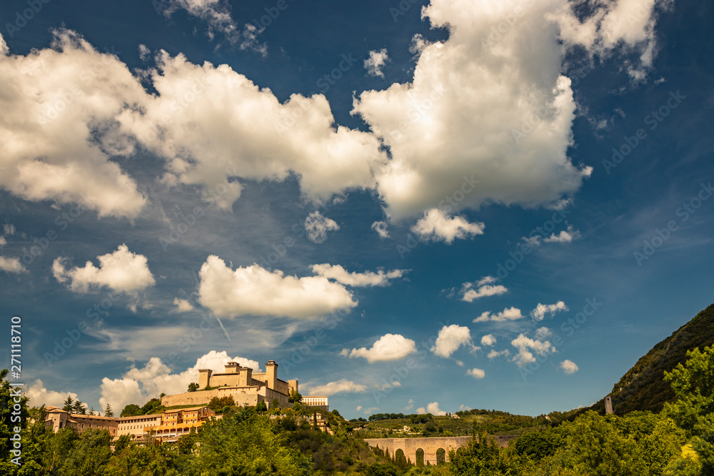 View of Spoleto, green mountains, blue sky with white clouds. The Rocca Albornoziana fortress illuminated by the sun in summer. The bridge of the towers, Roman aqueduct. Trees in the foreground