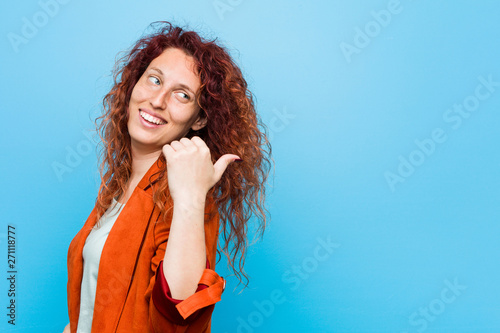 Young redhead elegant woman points with thumb finger away, laughing and carefree.