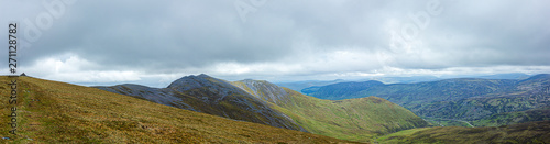 A panorama view of a Scottish mountain valley with a road and mountain range under a stormy huge white clouds sky