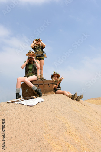 Young twin sisters and an adult girl dress up as explorers. They pose on a sand dune hill dressed with jungle hats and khaki safari clothes.