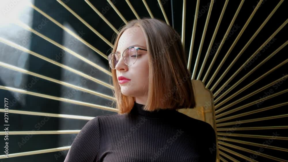 Attractive red-haired young woman with a short haircut in a square, wearing glasses and a brown turtleneck against a wall with a decor of golden rays. Daylight room. Attractive red-haired young woman with a short haircut in a square, wearing glasses and a brown turtleneck against a wall with a decor of golden rays. Daylight room.
