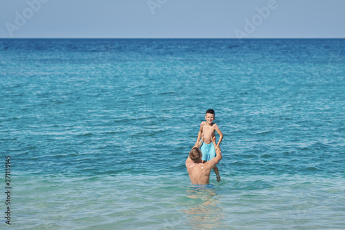 Man is holding son in his hands while standing in the ocean. They are enjoying their vacations, having fun and smiling.