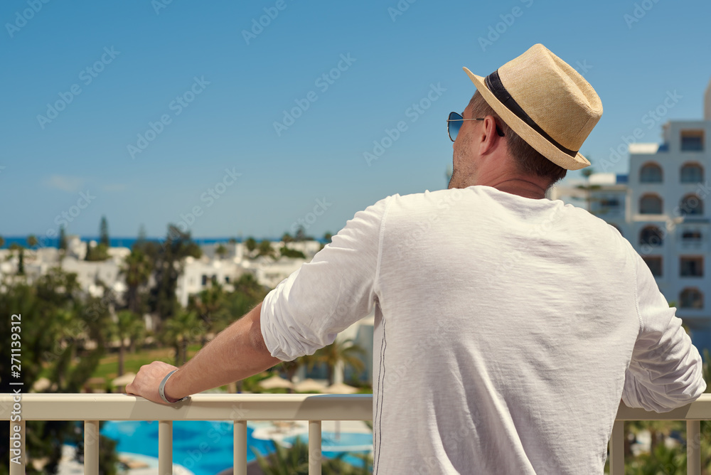 Handsome man in sunhat and white t-shirt is standing on the hotel’s balcony and looking towards blue sea. He is enjoying picturesque view.