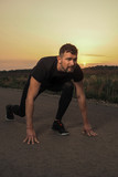 Mature fitness coach or runner in black sport joggers and tee stands in getting ready pose on a free countryside road in the evening time