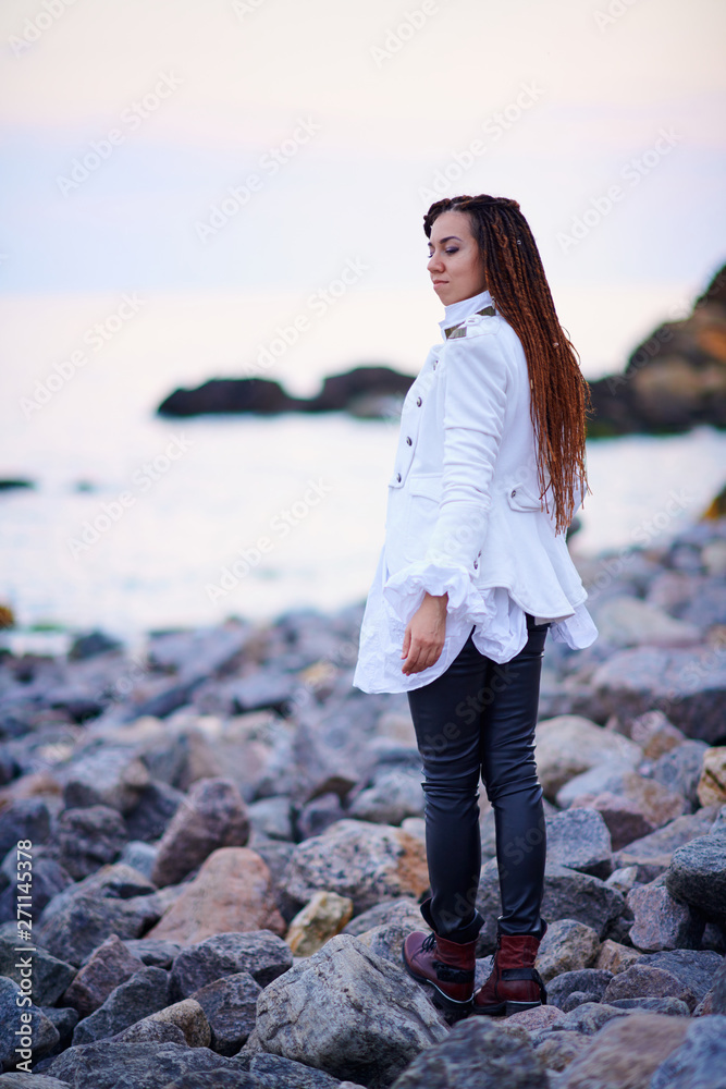 dreadlocks fashionable girl dressed in white jacket and black leather trousers posing near sea in the evening