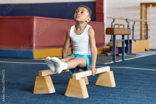Picture of adorable dark skinned little gymnast competing on parallel bars. Hardworking talented African child exercising at gym, doing acrobatic moves, demonstrating strength, agility and flexibility