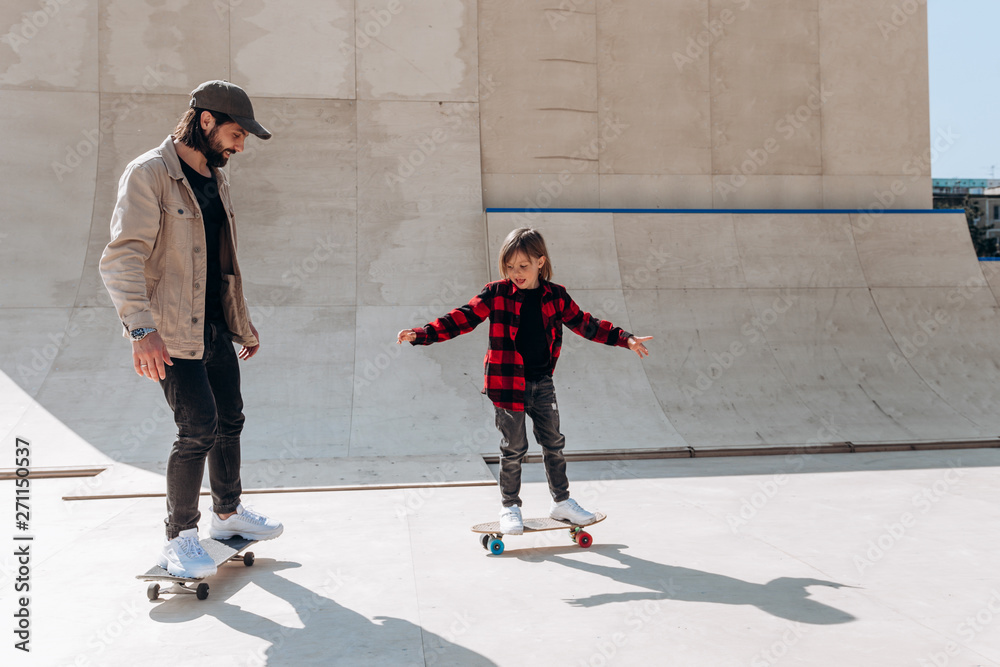 Obraz premium Ojciec i jego synek ubrani w zwykłe ciuchy jeżdżą na deskorolkach w skateparku ze zjeżdżalniami na zewnątrz w słoneczny dzień