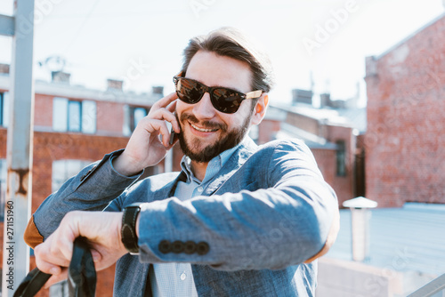 Handsome businessman wearing suit, stubble and sunglasses walking on the roof. Talking on the phone and watching his wristwatch same time