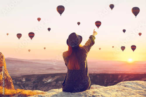 A woman alone unplugged sits on top of a mountain and admires the flight of hot air balloons in Cappadocia in Turkey. Digital detox and soul search