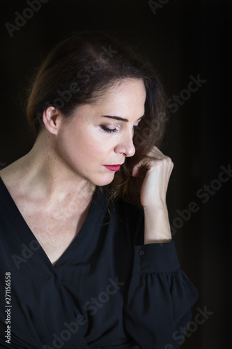 Portrait of a woman 48 years old, looking down smelling her wrist with closed eyes, isolated over black, low key lighting.