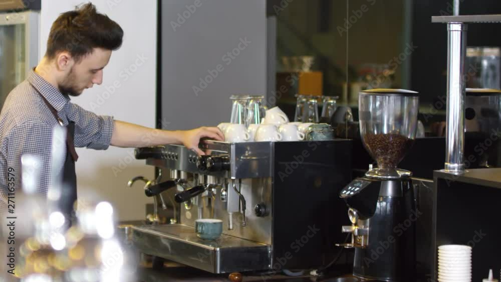 Waist-up shot of Caucasian male barista standing by coffee machine and waiting while double espresso is being brewed, then picking up ceramic cup and filling it with boiling water to make Americano Waist-up shot of Caucasian male barista standing by coffee machine and waiting while double espresso is being brewed, then picking up ceramic cup and filling it with boiling water to make Americano