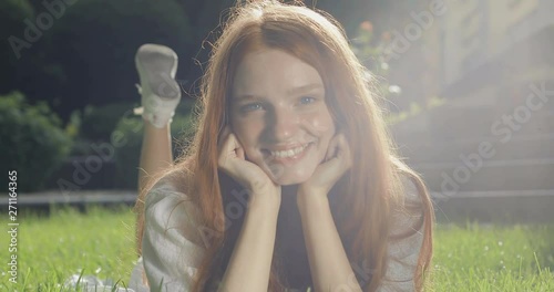 Cute red-haired girl smiling lying on the grass near the house. Young woman wiggles her legs and looks happy and fun. tracking shot. real time. Shot on Canon 1DX mark2 4K camera photo