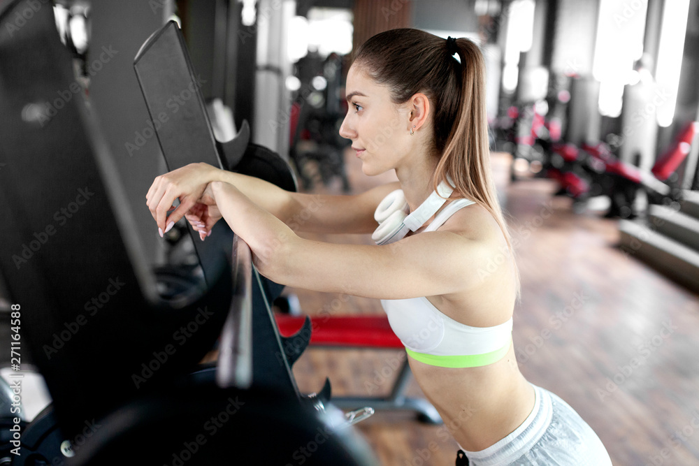 Fototapeta premium Closeup portrait of young pretty european fitness woman with headphones standing near the barbell at the gym. Breaking relax while exercise workout. Нealth and sport lifestyle. Athletic Body.