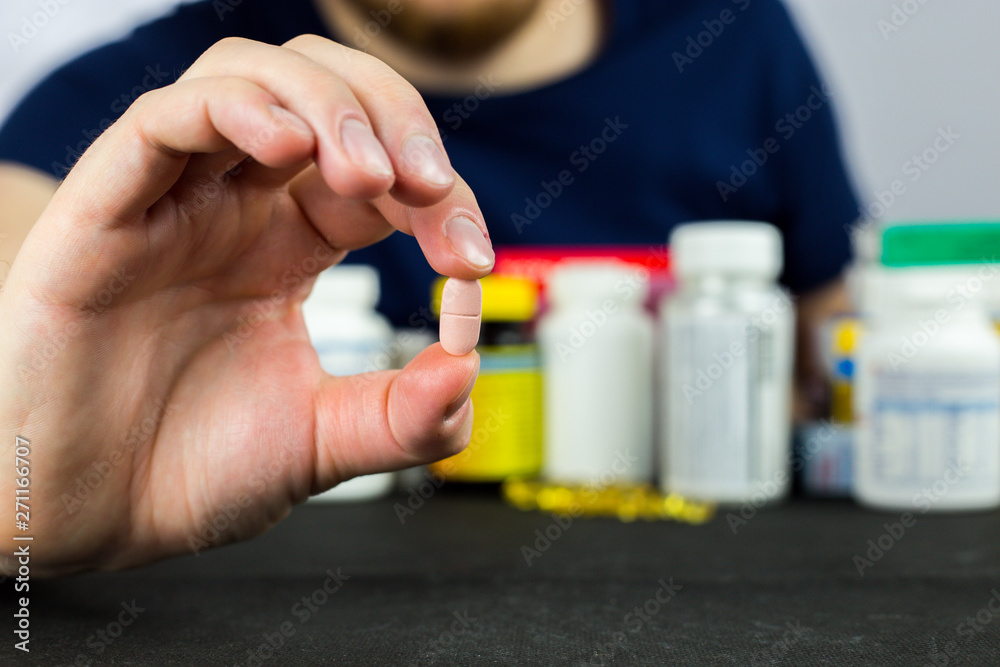 The person holds the pill between the fingers, offers the medicine, vitamins, in the background more pills, vitamins, medicines The person holds the pill between the fingers, offers the medicine, vitamins, in the background more pills, vitamins, medicines