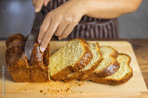 A person slicing a loaf of Brioche bread toast with a serrated knife on wood cutting board.